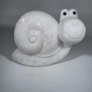 May include: A white, carved snail sculpture with a smiling face and black eyes. The snail's shell features a spiral design. The sculpture is made of a light-colored material and has a smooth, polished appearance. The snail is a whimsical decorative item.