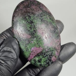 May include: An oval-shaped gemstone with a mix of colors, including deep green, dark purple, and black. The stone has a polished surface and a natural, textured appearance. The gemstone is held in a gloved hand.