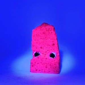 May include: A vibrant, neon pink rock formation with two dark, circular holes, set against a deep blue background. The porous texture of the rock is visible, and the lighting creates a glowing effect. The overall aesthetic is striking and unusual.