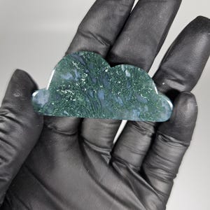 May include: A cloud-shaped decorative stone with a moss agate pattern. The stone features a mix of green, white, and blue hues, creating a natural, textured appearance. The cloud is held in a gloved hand, showcasing its size and shape.