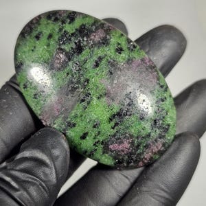 May include: An oval-shaped polished stone with a vibrant green base, speckled with black and hints of pink and purple. The stone is held in a black gloved hand, showcasing its natural colors and textures. A unique gemstone.
