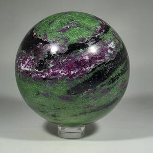 May include: A polished sphere of zoisite with ruby inclusions. The gemstone features a vibrant green base color with streaks of black and patches of deep magenta. The sphere is displayed on a clear acrylic stand, showcasing its natural beauty.