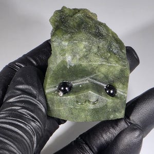 May include: A carved green stone sculpture with black bead eyes and a carved mustache. The stone has a rough, natural texture and is held in a black gloved hand. The sculpture has a whimsical, cartoonish appearance.