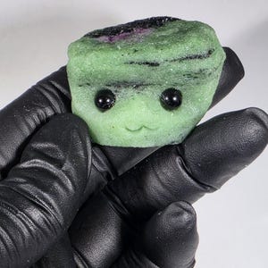 May include: A green, stone-like sculpture with black, beady eyes and a small, smiling mouth. The top of the sculpture has a dark purple and black stripe. The sculpture is held by a gloved hand.
