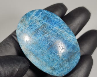 May include: A polished, oval-shaped blue apatite gemstone. The stone has a mottled appearance with varying shades of blue and subtle veining. The gemstone is held in a gloved hand.