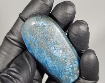 May include: A polished, oval-shaped blue gemstone with streaks of silver and brown. The stone is held in a black gloved hand against a gray background. The gemstone's surface has a shimmering, textured appearance.