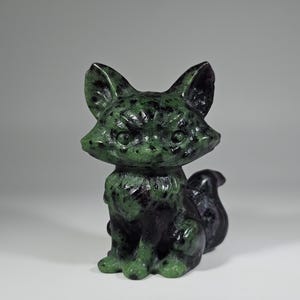 May include: A carved fox figurine in shades of green and black. The fox is sitting upright with its tail curled. The figurine has a textured surface and detailed features, including pointed ears and a fluffy collar.