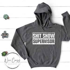 May include: A dark grey hooded sweatshirt with the text "SHIT SHOW SUPERVISOR" printed in white on the front.