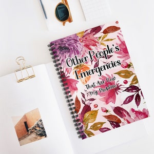 以下が含まれることがあります： ピンク、紫、バーガンディの色合いの花柄デザインの、スパイラル綴じノート。表紙には「Other People's Emergencies That Are Now My Problem」のテキストが書かれています。ノートは白い表面に開かれています。