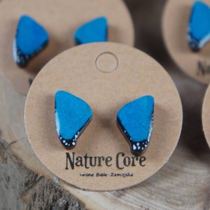 Op de afbeelding: Een paar blauwe vlinder-vormige oorbellen met zwarte en witte accenten. De oorbellen worden getoond op een bruin houten oppervlak met een label waarop "Nature Core" en "Iwona Biela - Zamojska" staat.