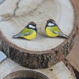 May include: Two miniature bird figurines with yellow, green, and brown coloring. The birds are perched on a wooden slice with a natural bark edge. The birds have a detailed design and are set against a blurred background.