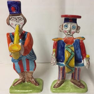 May include: Two ceramic figurines of musicians. The one on the left is playing a yellow saxophone and is wearing a blue hat, red and blue striped pants, and a red shirt. The one on the right is playing a drum and is wearing a red hat, blue and red striped pants, and a red shirt.