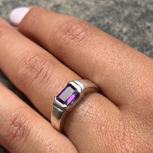 May include: A silver ring with a purple gemstone set in a rectangular shape. The ring has a simple design with a smooth band.