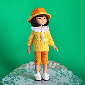 Puede incluir: Una muñeca con un suéter de punto amarillo con un cuello de encaje blanco, pantalones cortos de punto naranja y un sombrero naranja y marrón. La muñeca tiene el pelo negro y los ojos marrones. La muñeca está de pie sobre un encaje blanco.