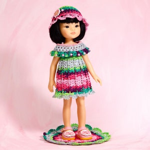 Puede incluir: Una muñeca de pelo negro con un vestido de ganchillo multicolor en tonos rosa, morado, verde y gris. La muñeca lleva un sombrero de ganchillo a juego con una pequeña flor y zapatos rosas. La muñeca está de pie sobre una alfombra de ganchillo.