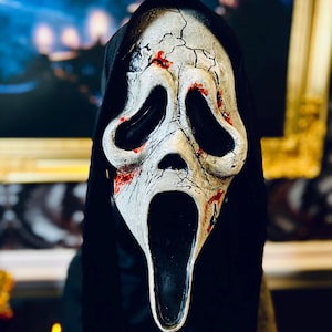 May include: A white, elongated ghost mask with a distressed, cracked texture and red blood-like splatters. The mask features large, hollow black eye sockets and an open, elongated mouth, attached to a black fabric hood that drapes over the sides.