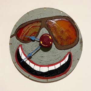Pode incluir: Um relógio de parede redondo e caprichoso com um design de rosto pintado. O relógio apresenta óculos de sol castanhos, um nariz vermelho e uma boca larga e sorridente. Os ponteiros do relógio são azuis e o mostrador é cinzento com marcas amarelas.
