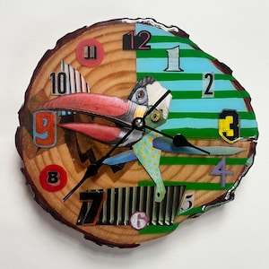 Pode incluir: Um relógio de parede único feito de uma seção transversal de um tronco de árvore. O mostrador do relógio apresenta um design de pássaro colorido com detalhes em vermelho, azul e amarelo. Os ponteiros do relógio são pretos e os números são de várias cores.