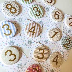 May include: A set of 10 round fabric buttons with different patterns and numbers from 0 to 8. The buttons are on white cards with colourful polka dots and the text "May Cat Hooray!" printed on them.