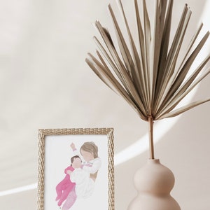 K&ouml;nnte beinhalten: Eine gerahmte Aquarellillustration einer Frau, die ein Baby h&auml;lt. Die Frau tr&auml;gt ein wei&szlig;es Hemd und das Baby tr&auml;gt ein rosa Outfit. Der Rahmen ist golden und hat ein gewebtes Muster.