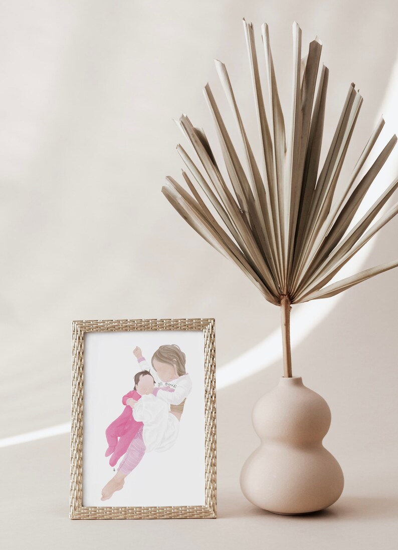 K&ouml;nnte beinhalten: Eine gerahmte Aquarellillustration einer Frau, die ein Baby h&auml;lt. Die Frau tr&auml;gt ein wei&szlig;es Hemd und das Baby tr&auml;gt ein rosa Outfit. Der Rahmen ist golden und hat ein gewebtes Muster.