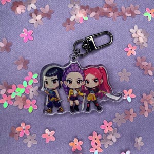 May include: A clear acrylic keychain featuring three anime-style characters with vibrant hair colors: black, purple, and red. The keychain has a silver clasp and is surrounded by pink and iridescent flower-shaped confetti.