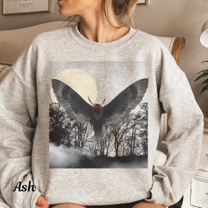 May include: A light gray sweatshirt featuring a graphic of a black moth with red eyes and a large moon in the background. The moth is set against a backdrop of a forest with bare trees. The word "Ash" is printed on the bottom left.