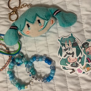 Op de afbeelding: Een pluche sleutelhanger van een personage met turkoois haar, twee elastische armbanden, twee kralenarmbanden in blauwtinten met letterkralen die I LOVE MIKU spellen, en drie stickers met een anime-personage met turkoois haar in diverse poses.