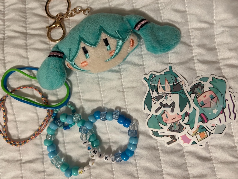 Op de afbeelding: Een pluche sleutelhanger van een personage met turkoois haar, twee elastische armbanden, twee kralenarmbanden in blauwtinten met letterkralen die I LOVE MIKU spellen, en drie stickers met een anime-personage met turkoois haar in diverse poses.