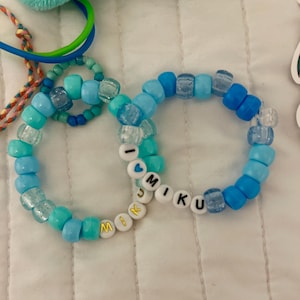 Op de afbeelding: Twee ronde armbanden gemaakt van diverse tinten blauwe plastic kralen. E&eacute;n armband bevat witte letterkralen die I HEART MIKU spellen met een blauw hartje, terwijl de andere witte kralen met gouden letters bevat die MIKU spellen.