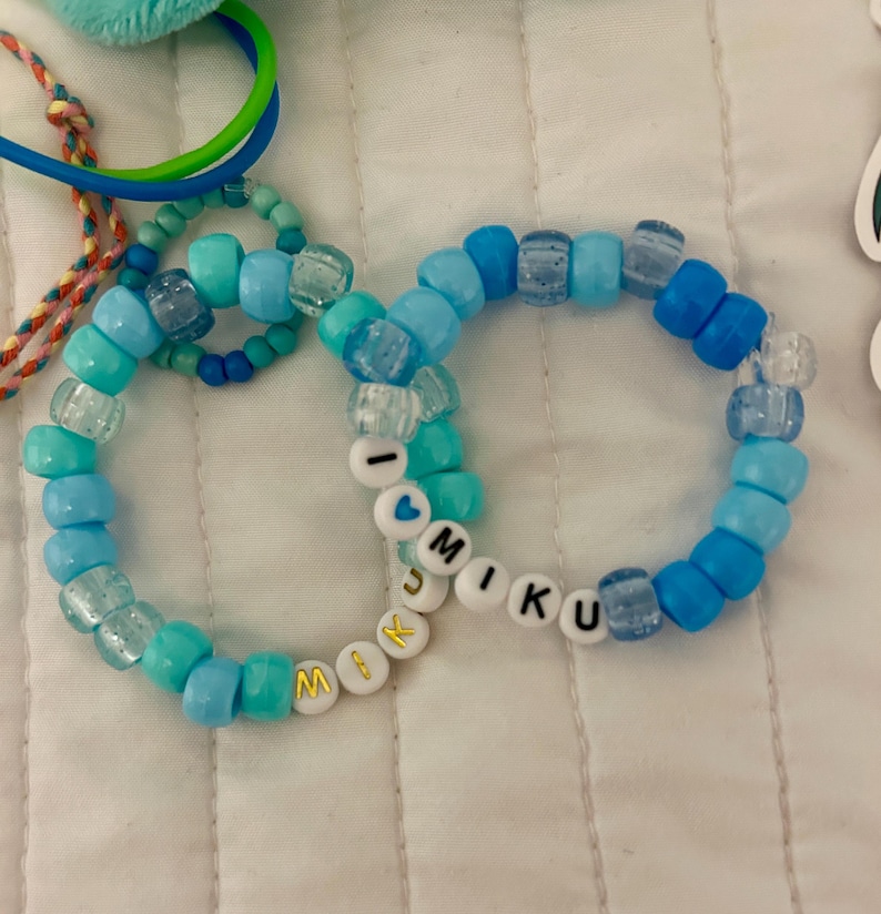 Op de afbeelding: Twee ronde armbanden gemaakt van diverse tinten blauwe plastic kralen. E&eacute;n armband bevat witte letterkralen die I HEART MIKU spellen met een blauw hartje, terwijl de andere witte kralen met gouden letters bevat die MIKU spellen.