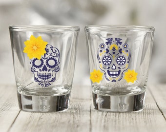 May include: Two clear shot glasses with Day of the Dead skull designs. One skull has a yellow flower and the other has yellow flowers and decorative elements. The skulls are navy blue and white.