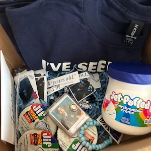 Op de afbeelding: Een marineblauw Gildan t-shirt met tekst, een pot Jet-Puffed marshmallowcr&egrave;me, twee Jif To Go pindakaascups, een kleine fotosleutelhanger, een lichtblauwe kralenarmband en diverse papieren uitknipsels in een kartonnen doos.