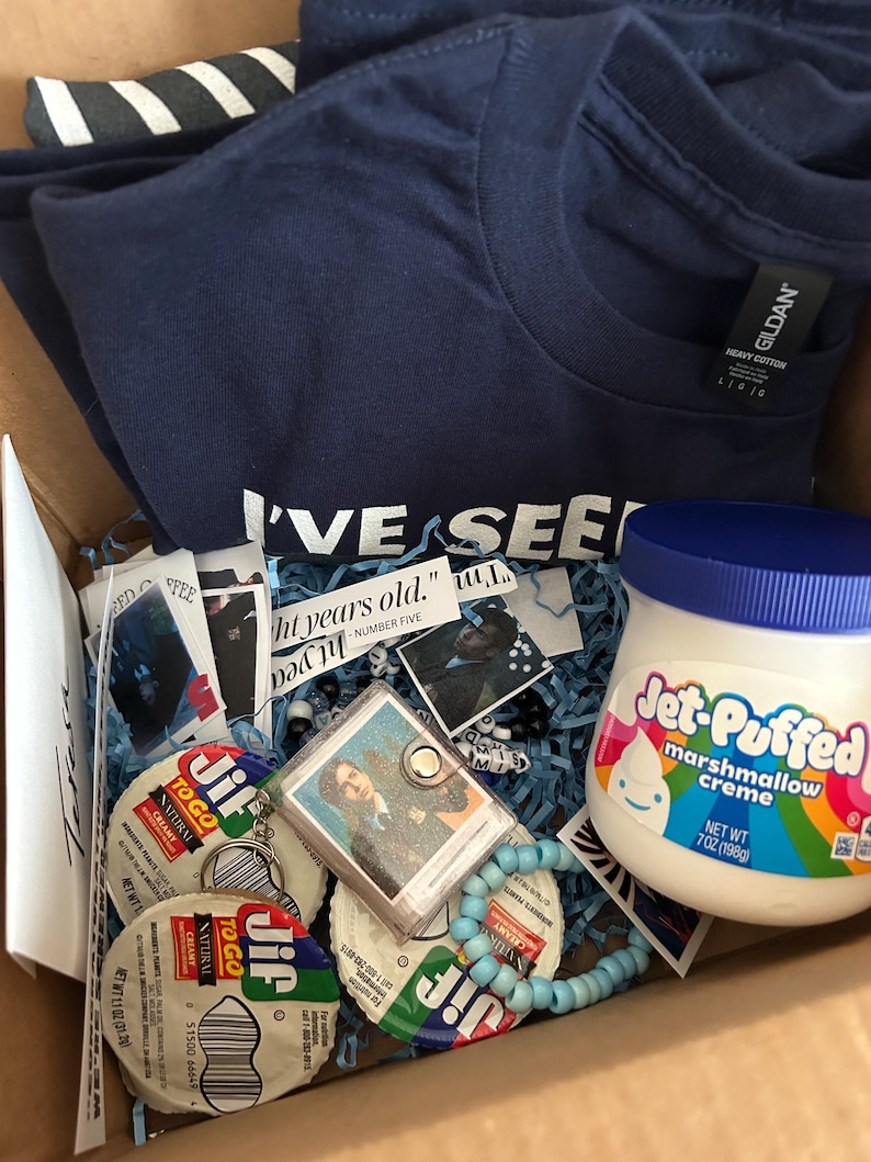Op de afbeelding: Een marineblauw Gildan t-shirt met tekst, een pot Jet-Puffed marshmallowcr&egrave;me, twee Jif To Go pindakaascups, een kleine fotosleutelhanger, een lichtblauwe kralenarmband en diverse papieren uitknipsels in een kartonnen doos.