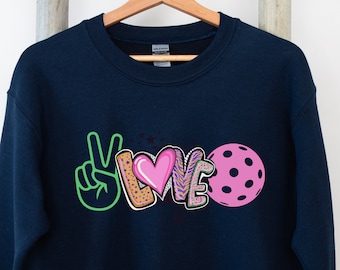 May include: Navy blue sweatshirt with a graphic design of the word "LOVE" in colorful, stylized letters, a peace sign, and a pink pickleball.