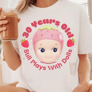 May include: White t-shirt with a pink strawberry graphic and the text "30 Years Old Still Plays With Dolls".