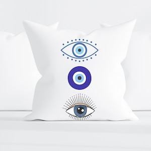 May include: White pillow with three blue evil eye designs. The top design is a stylized eye with a blue iris and pupil, surrounded by a dotted circle. The middle design is a solid blue circle with a smaller white circle inside. The bottom design is a stylized eye with a black pupil and a blue iris, surrounded by radiating lines.
