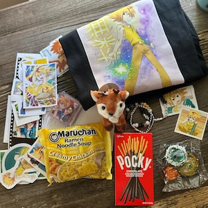 Op de afbeelding: Een zwart T-shirt met een anime-personage met geel haar, een kleine pluche ree, een pak Maruchan Creamy Chicken Ramen, een doos Pocky chocoladesticks, een kralenarmband, diverse anime-stickers en een plastic speelgoedonderdeel.