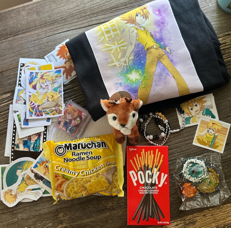 Op de afbeelding: Een zwart T-shirt met een anime-personage met geel haar, een kleine pluche ree, een pak Maruchan Creamy Chicken Ramen, een doos Pocky chocoladesticks, een kralenarmband, diverse anime-stickers en een plastic speelgoedonderdeel.