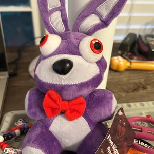 Op de afbeelding: Een paarse pluche konijnknuffel met lange oren, grote witte ogen met rode pupillen, een zwarte neus en een felrode vlinderdas. Een label aan de knuffel bevat de tekst Five Nights at Freddy's en Collectible Plush in witte letters.