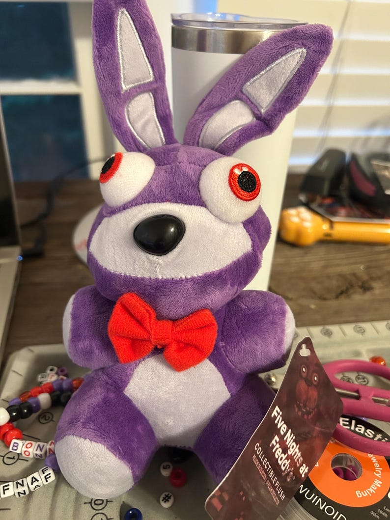 Op de afbeelding: Een paarse pluche konijnknuffel met lange oren, grote witte ogen met rode pupillen, een zwarte neus en een felrode vlinderdas. Een label aan de knuffel bevat de tekst Five Nights at Freddy's en Collectible Plush in witte letters.