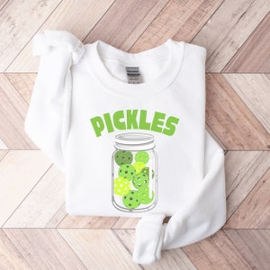 May include: White sweatshirt with the text "PICKLES" in green letters above a drawing of a glass jar filled with green and yellow pickleball balls.