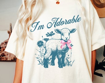 Peut inclure: Un t-shirt blanc avec un graphique bleu d'un agneau avec un nœud rose. Le texte "I'm Adorable" est écrit en cursive bleue au-dessus de l'agneau.