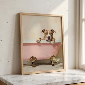 May include: A framed wall art print featuring a tan pit bull dog resting its front paws on the edge of a pink clawfoot bathtub. The tub has gold-coloured feet, and a glass of martini with a lime garnish sits on the floor beside the bathtub.