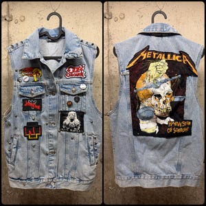 May include: Light blue denim vest adorned with patches featuring band logos and studded accents. The back displays a large Metallica patch with a skull graphic. The vest is hanging on a black hanger.