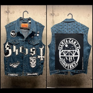 Op de afbeelding: Een denim vest versierd met patches en studs. De voorkant heeft patches met het woord "Ghost", een schedel en andere ontwerpen. De achterkant heeft een grote patch met de tekst "Santa Carla Vampires". Het vest is versierd met metalen studs.