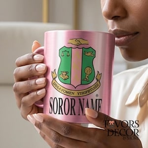 Puede incluir: Una taza metálica rosa con un escudo verde y rosa y el texto "SOROR NAME". La taza está sujeta por una persona. La taza tiene un asa en el lado izquierdo y las palabras "FAVORS DECOR & MORE" están en la esquina inferior derecha.