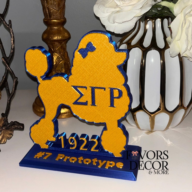 May include: A yellow and blue poodle-shaped decorative item. The poodle has a blue bow and the Greek letters &Sigma;&Gamma;&Rho;. The base reads "1922" and "#7 Prototype". The item is a decorative piece.