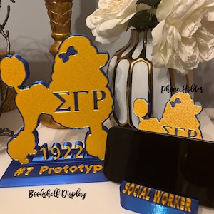 May include: A gold and blue poodle-shaped phone holder with the Greek letters &Sigma;&Gamma;&Rho; and the year 1922. The base reads "#7 Prototype" and "Bookshelf Display". A smaller holder reads "Social Worker". The words "Phone Holder" are written on the image.