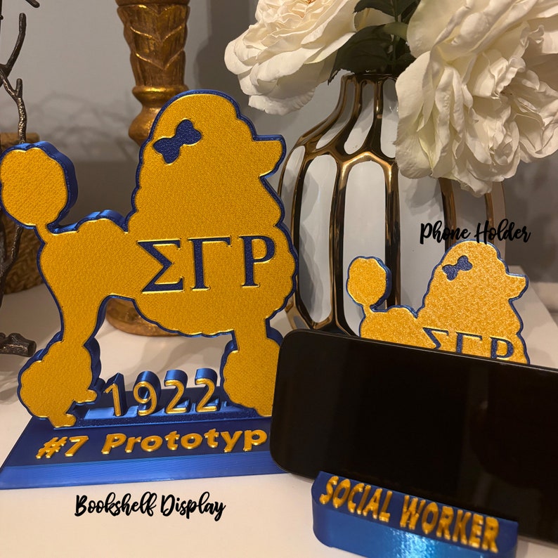 May include: A gold and blue poodle-shaped phone holder with the Greek letters &Sigma;&Gamma;&Rho; and the year 1922. The base reads "#7 Prototype" and "Bookshelf Display". A smaller holder reads "Social Worker". The words "Phone Holder" are written on the image.